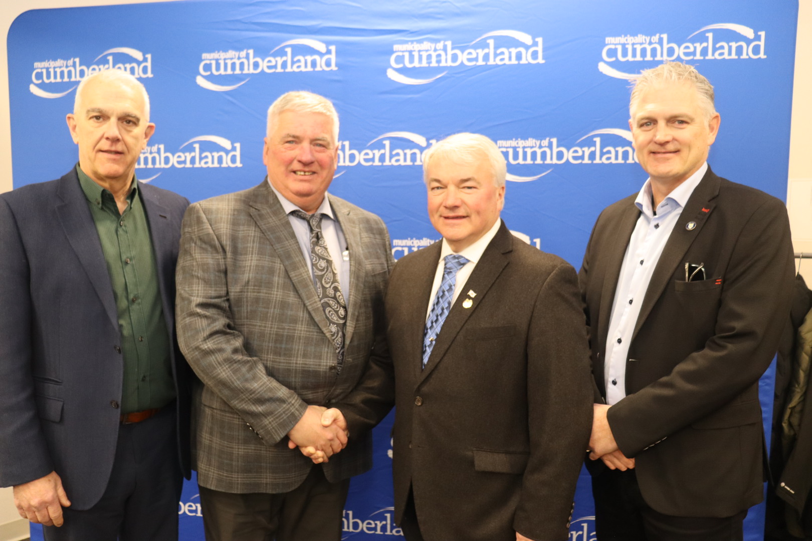 Four men stand in front of a banner with two men in the middle shaking hands.