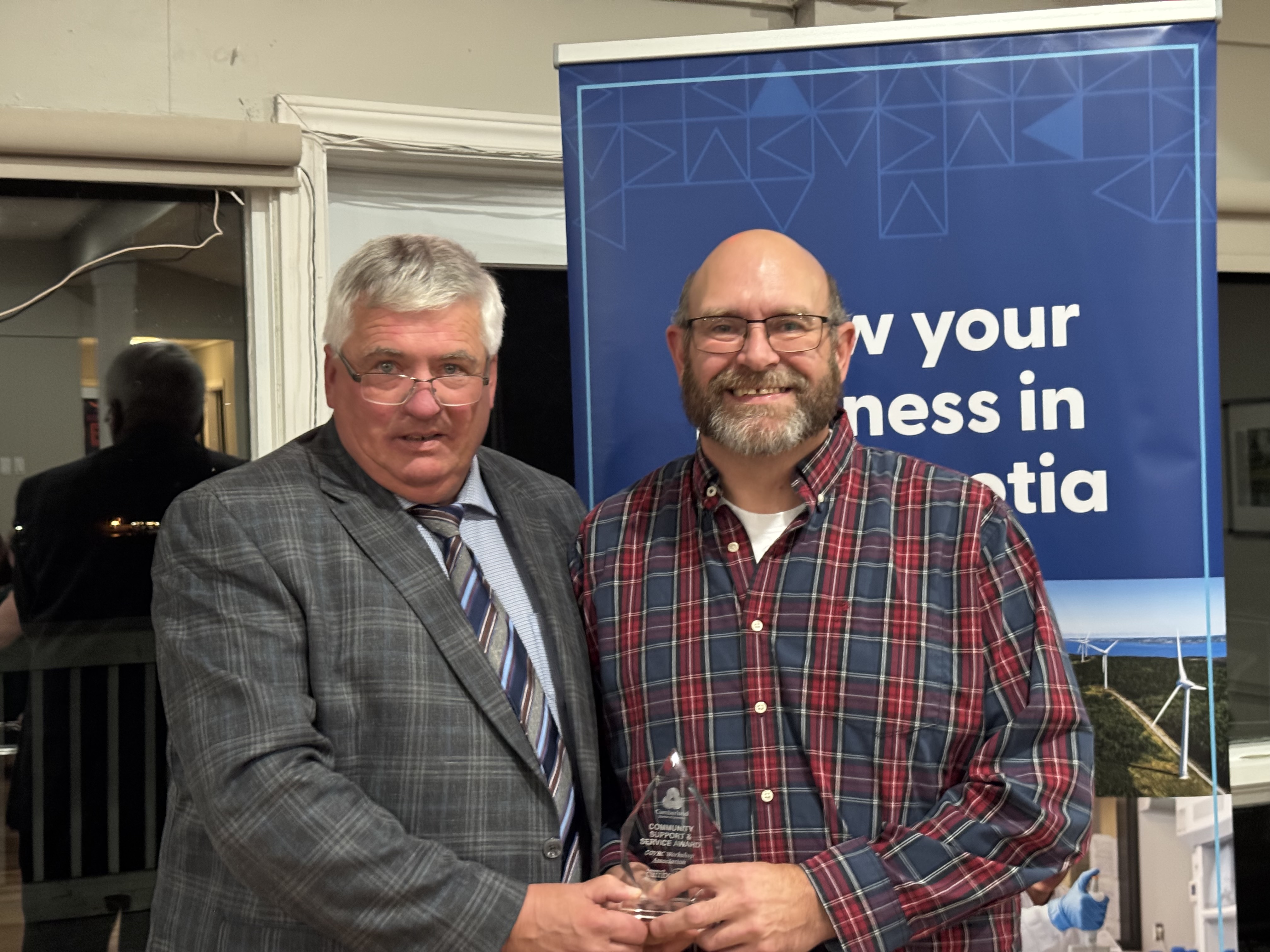 One man in a suit presents an award to another person with a tartan shirt while standing in front of a banner.