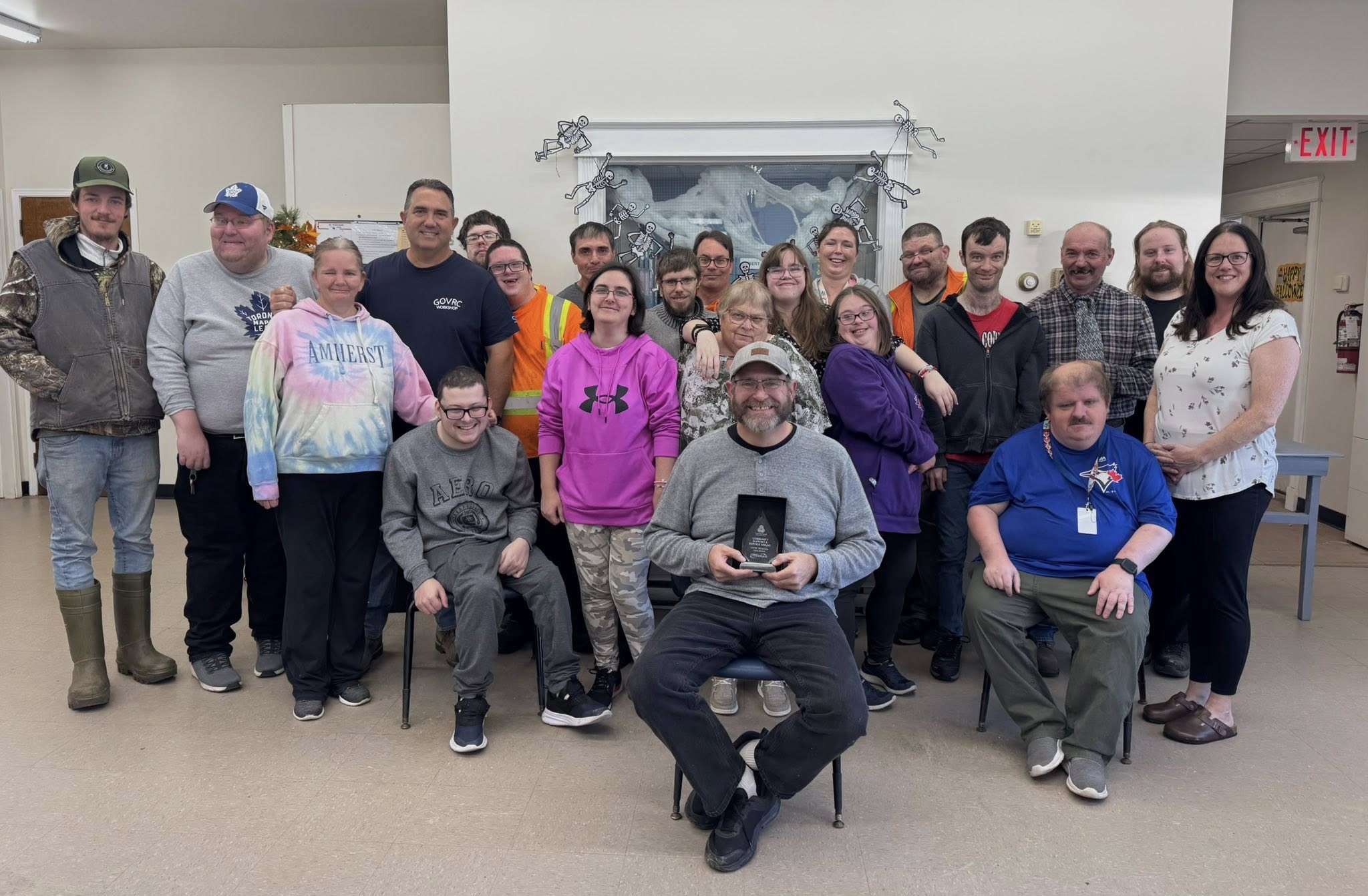 A group of people pose for a photo while a man is seated in the front with a trophy.
