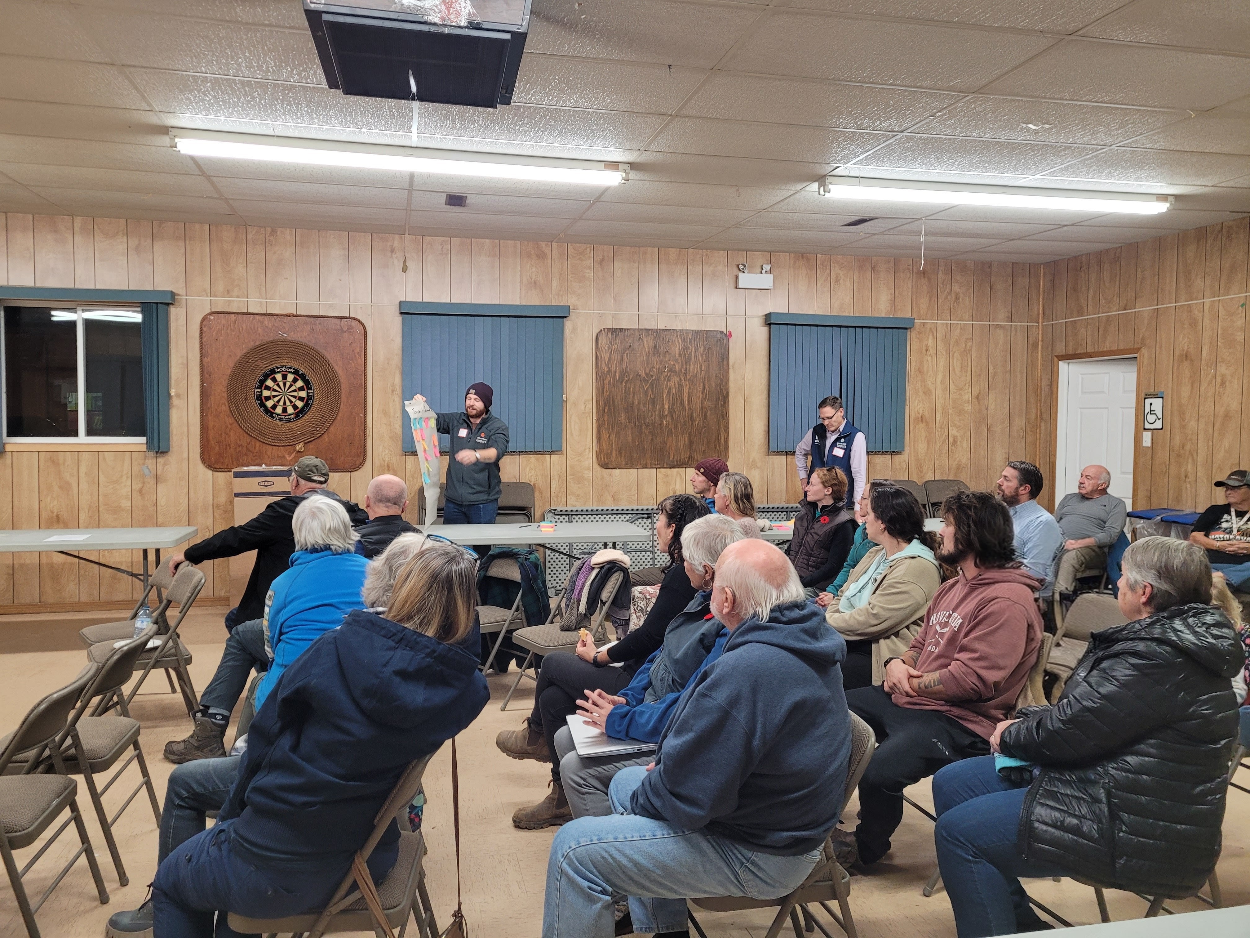 A group of people sit in chairs and watch two people at the front of the room give a presentation about the Cliffs of Fundy Global Geopark.