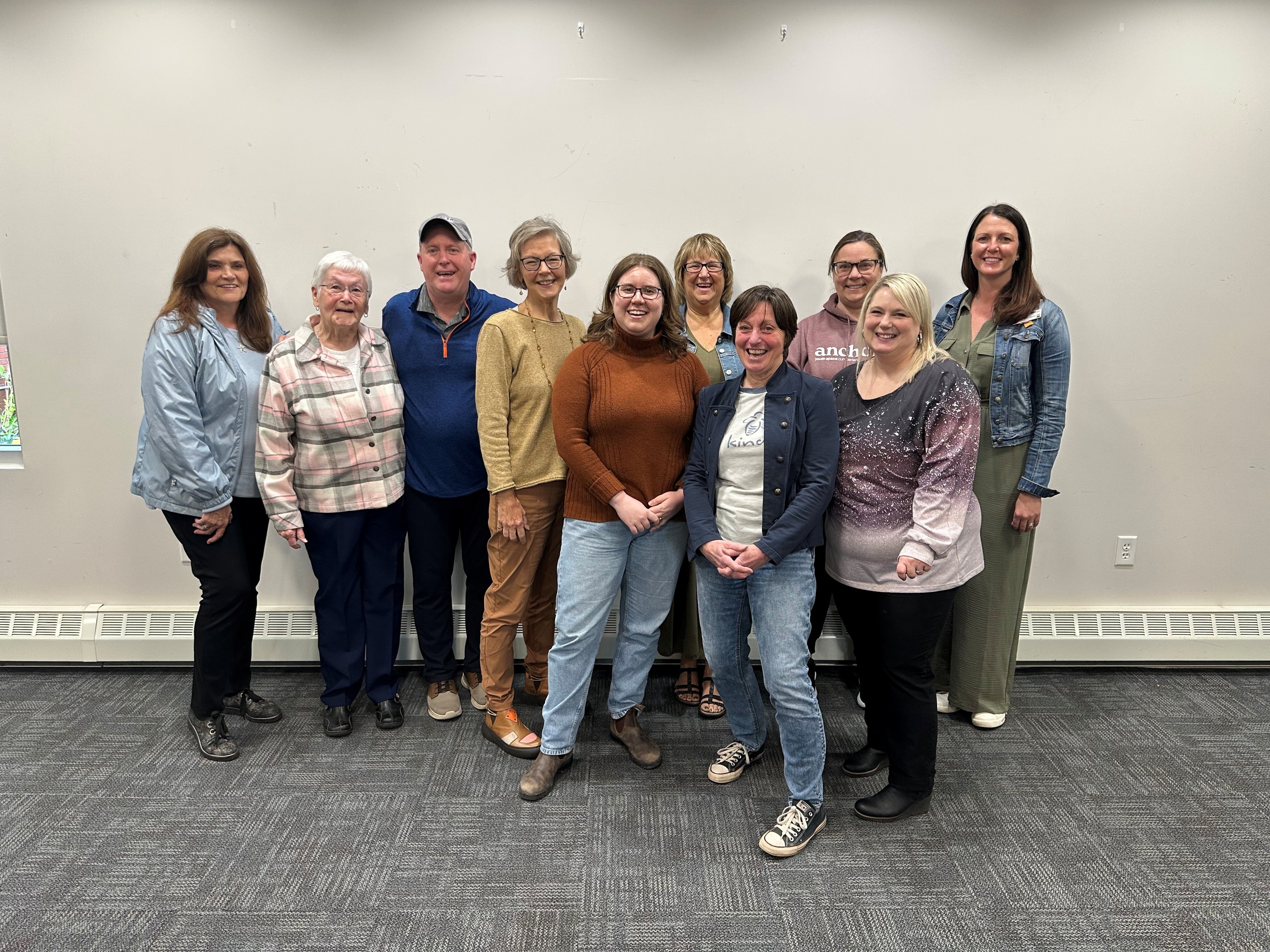 Nine women and one man stand in front of a white wall. 