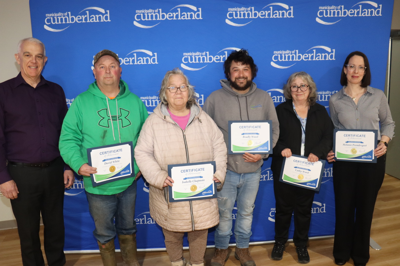 Three men and three women stand in front of a backdrop with the Municipality of Cumberland logo on it. Five people are holding certificates.
