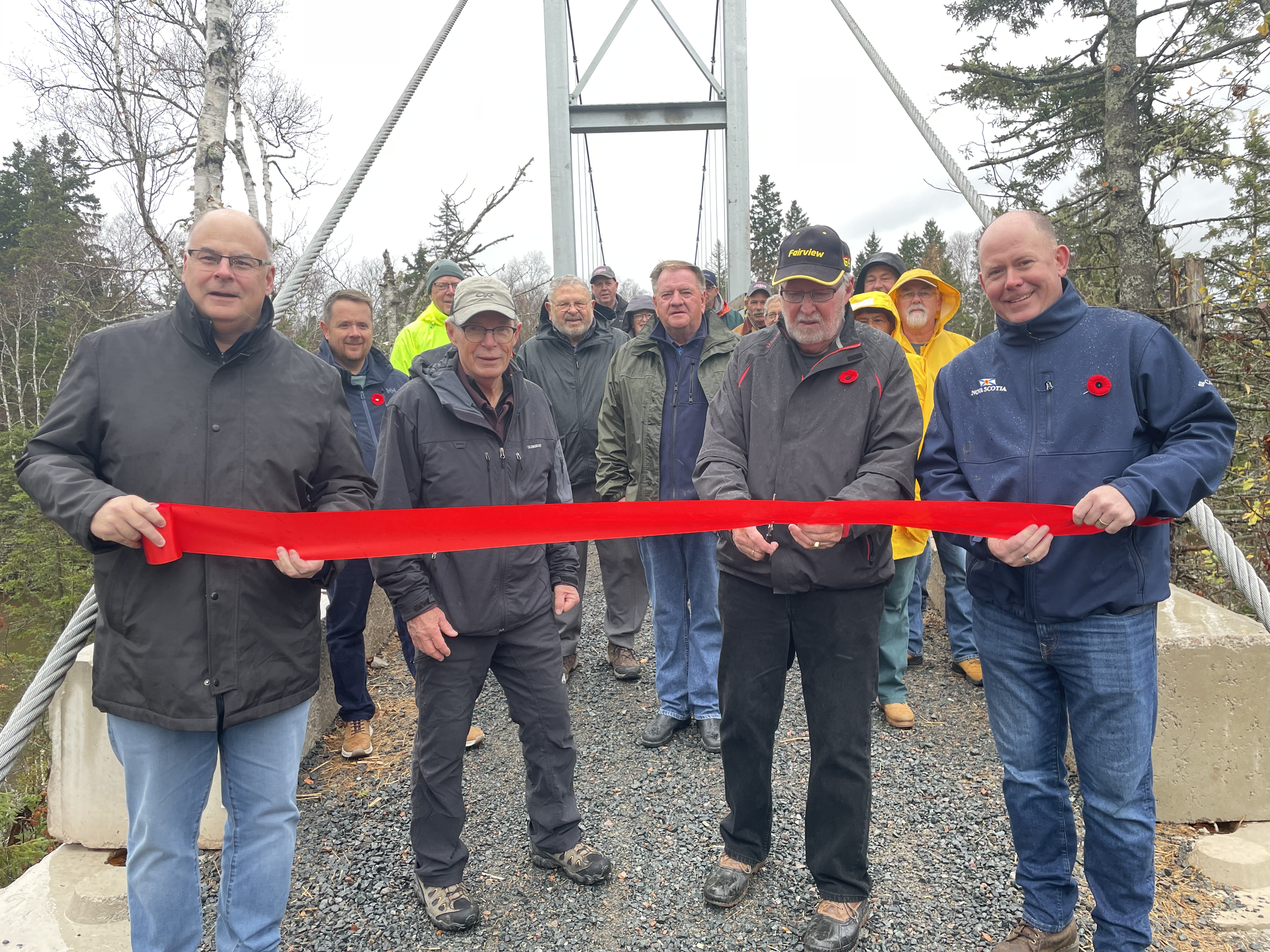 Four men stand in front of a ribbon while one of them prepares to cut it. A group of men and women are in the background.