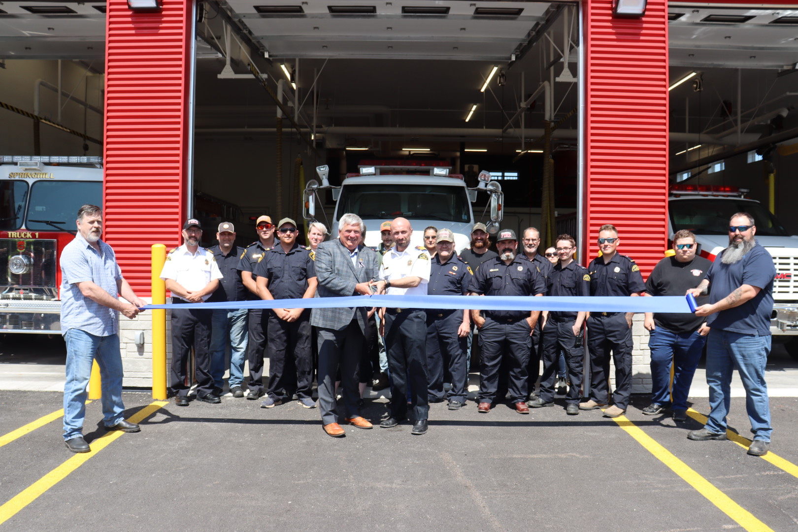 A group of people stand before a ribbon in front of a large building containing firefighting vehicles