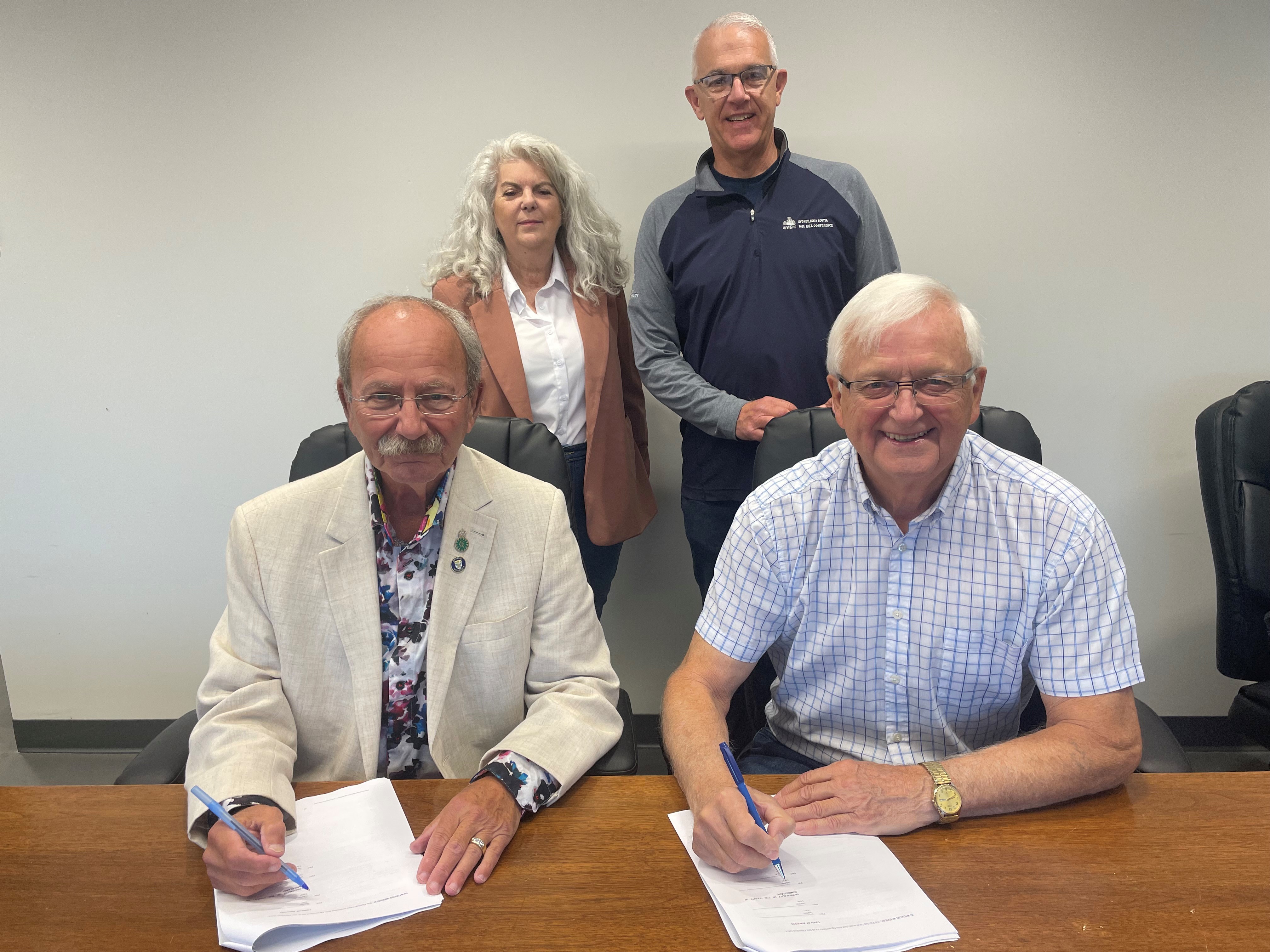 Two men are seated at a table signing a document while a woman and a man stand behind.