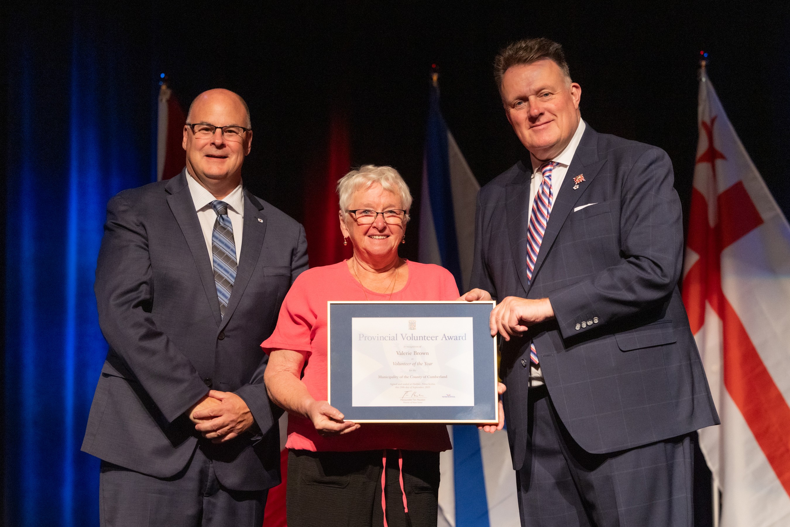 Two men present a provincial volunteer award to a woman on a stage with flags in the background.
